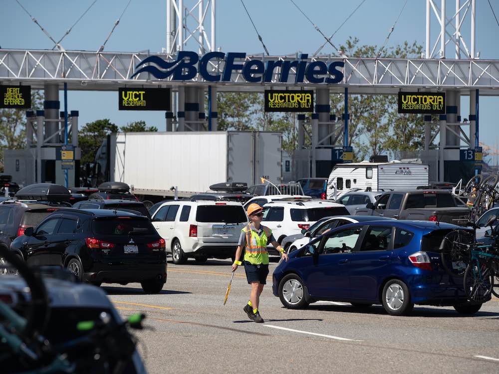 Vehicles are lined up outside a ferry terminal. A worker holds a sign for directing traffic. The fare booths are in the background.