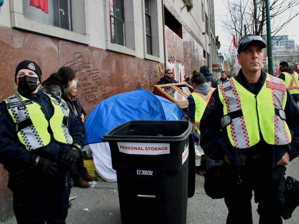 Two VPD officers, in black clothes and yellow reflective vests, look down the street. Between them is a black garbage bin labelled "Personal Storage." Behind them is a tent covered with a blue tarp and more people and possessions.