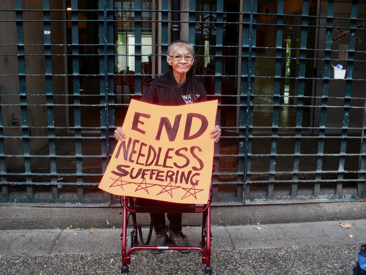 A woman with short hair and a walker stands in front of a neglected iron grating holding an orange hand-lettered sign that says "End needless suffering."