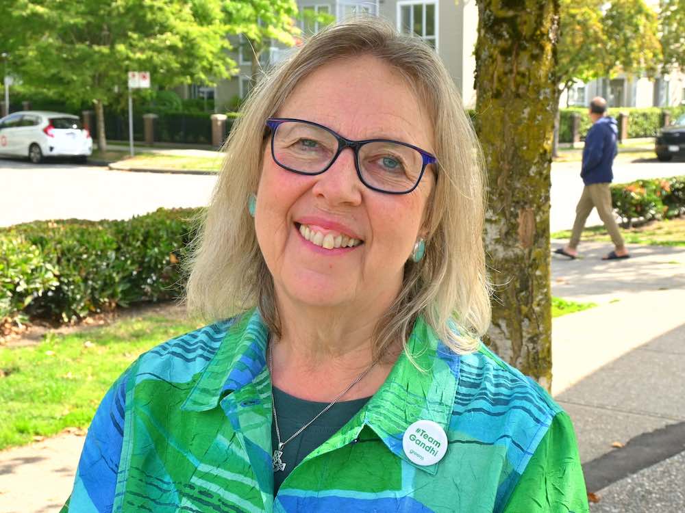 Elizabeth May stands outside wearing glasses and a jaunty blue and green collared silk shirt. Her blond hair comes to her shoulders. She smiles at the camera.