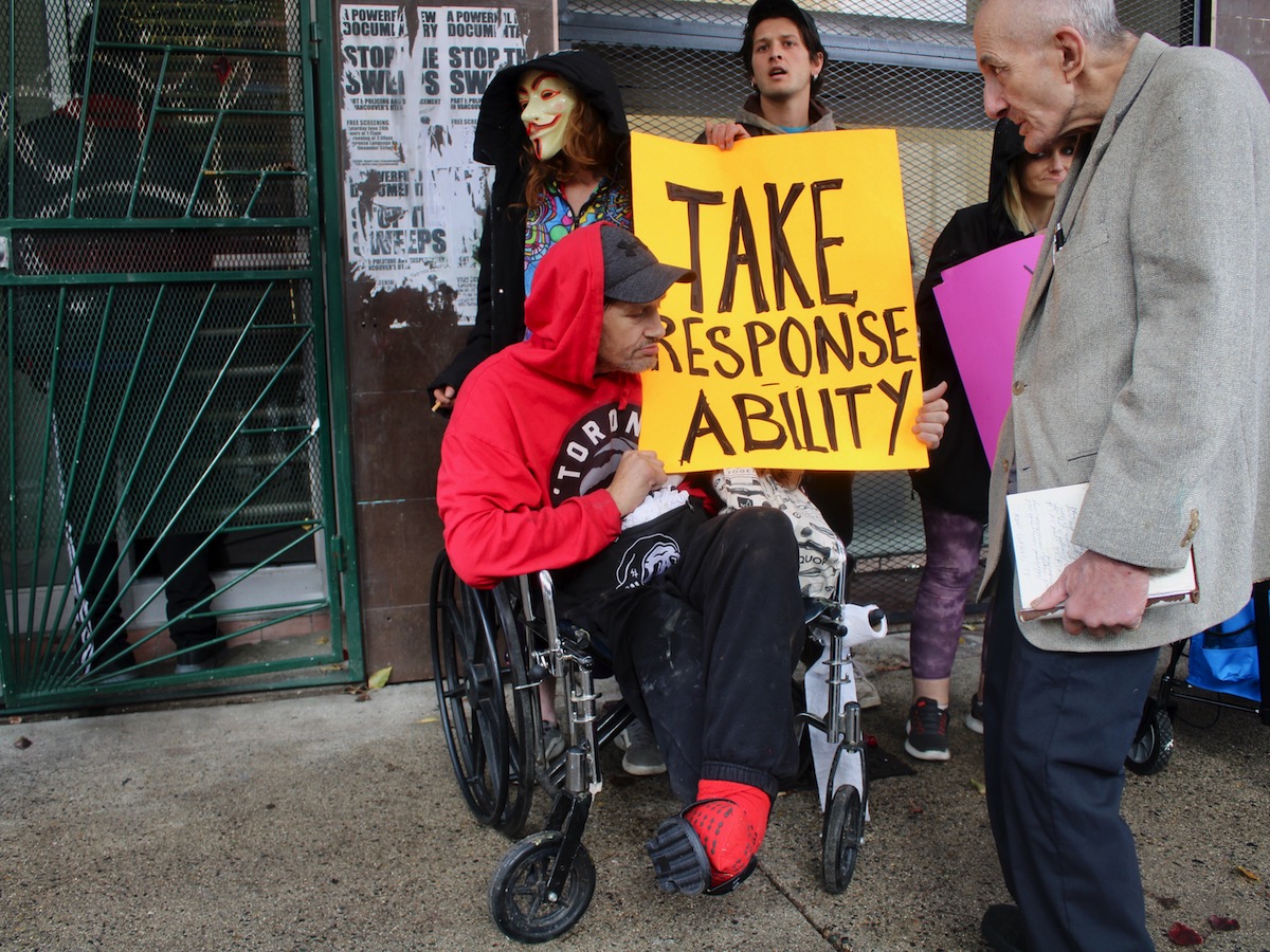 Five people are in front of a neglected hotel facade, including a man in a red hoodie in a wheelchair, with one leg, holding a sign that says "Take Response Ability." The group includes a person wearing a Guy Fawkes mask.