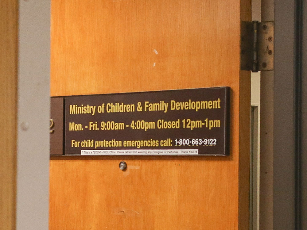 A wooden office door has a brown plastic sign that reads 'Ministry of Children & Family Development,' with opening times and a number to call for child protection emergencies. It appears as though it is from the '60s or '70s.
