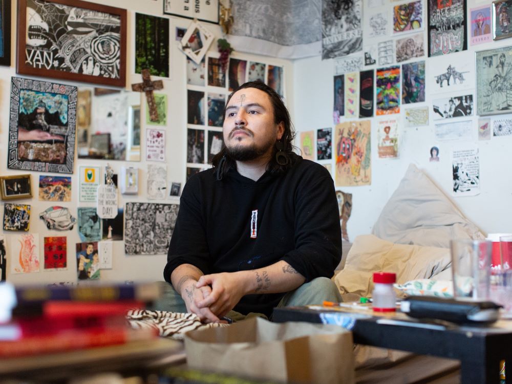 An Indigenous person with light skin, dark hair and a beard sits on a bed. They have a tattoo on their forehead and are wearing earrings. The walls behind them are densely packed with art.