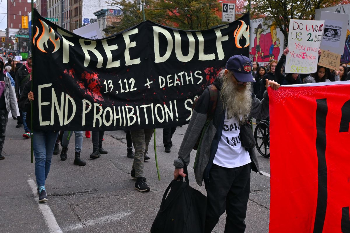 A banner at a rally reads, ‘FREE DULF. 13,112+ deaths. End Prohibition!’ It is covered with red handprints.