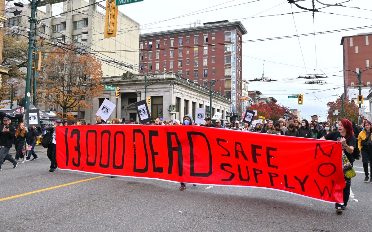 The rally for DULF moves through the downtown eastside. A group of people hold a banner that reads, ‘13,000 DEAD. SAFE SUPPLY NOW.’