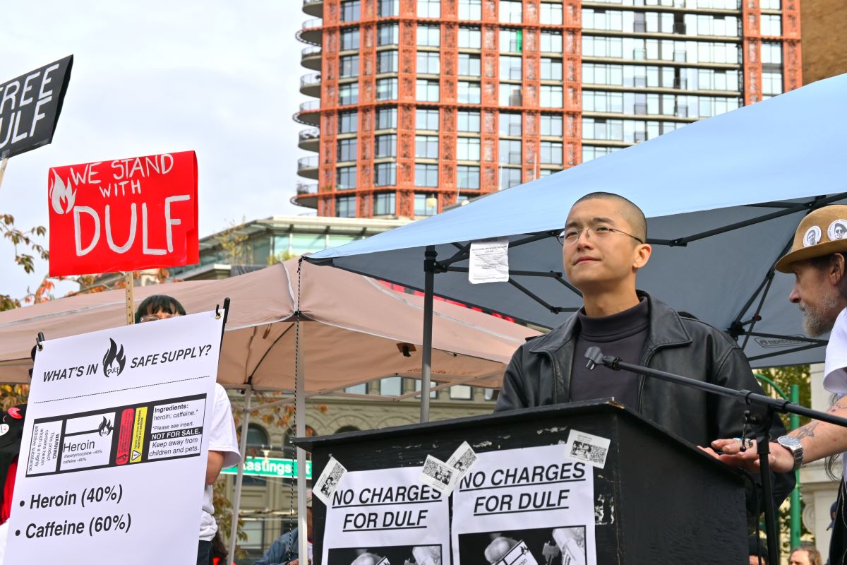 Vince Tao, a man with a shaved head and olive skin tone, stands at a podium at Victory Square in Vancouver. Posters taped to the podium read, ‘No Charges for DULF.’