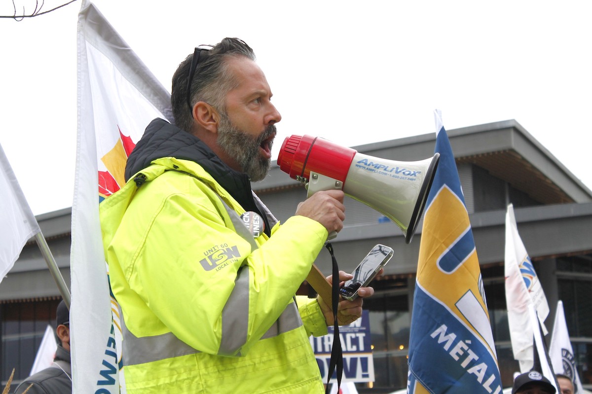 A dark-haired man with a beard, wearing a yellow safety jacket, speaks into a megaphone.