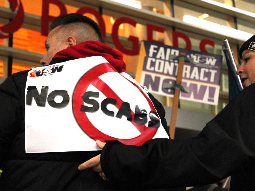 A man with short hair wears a sign saying 'No Scabs' on his back. The Rogers logo is visible behind him on an office building.