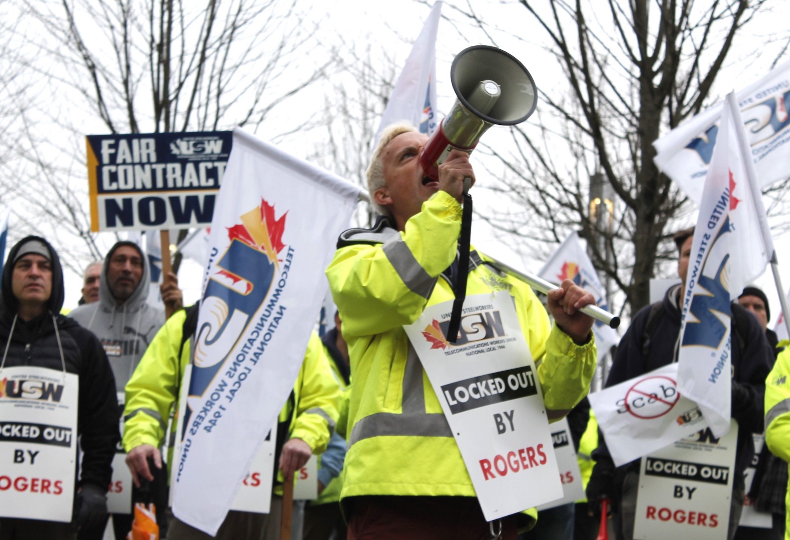 A white man with light-coloured hair speaks into a megaphone on a grey fall day. Behind him other men are lined up wearing signs that say 'Locked Out.'