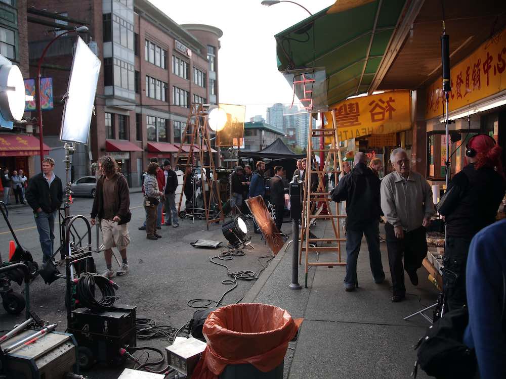 About 20 people work on a busy film shoot in Chinatown. Bright lights and reflectors are aimed at a storefront, where more film staff are at work.