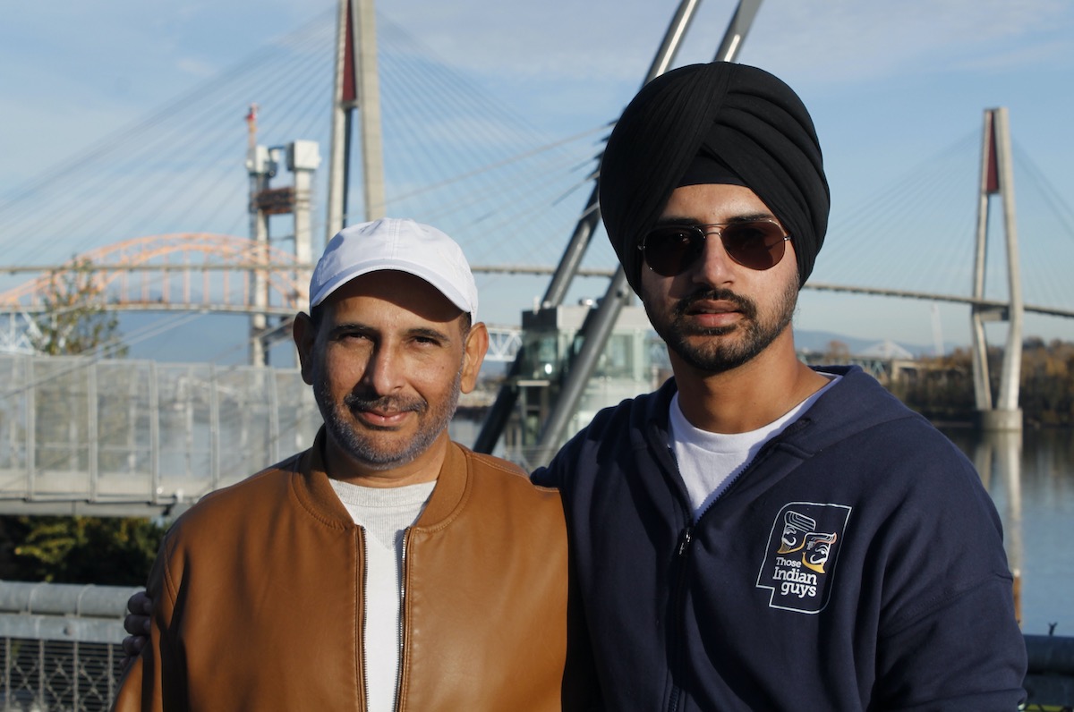 Two Indo-Canadian men look at the camera outdoors. The one on the left is wearing a white T-shirt and ball cap and light brown leather jacket and has a short beard. The one on the right is wearing a turban, sunglasses and a white T-shirt and blue jacket.