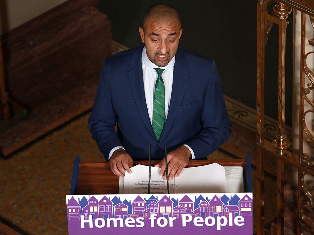 An Indo-Canadian man, balding and with a goatee, stands at a podium with a sign saying 'Homes for People.' He wears a white shirt, blue suit and green striped tie.