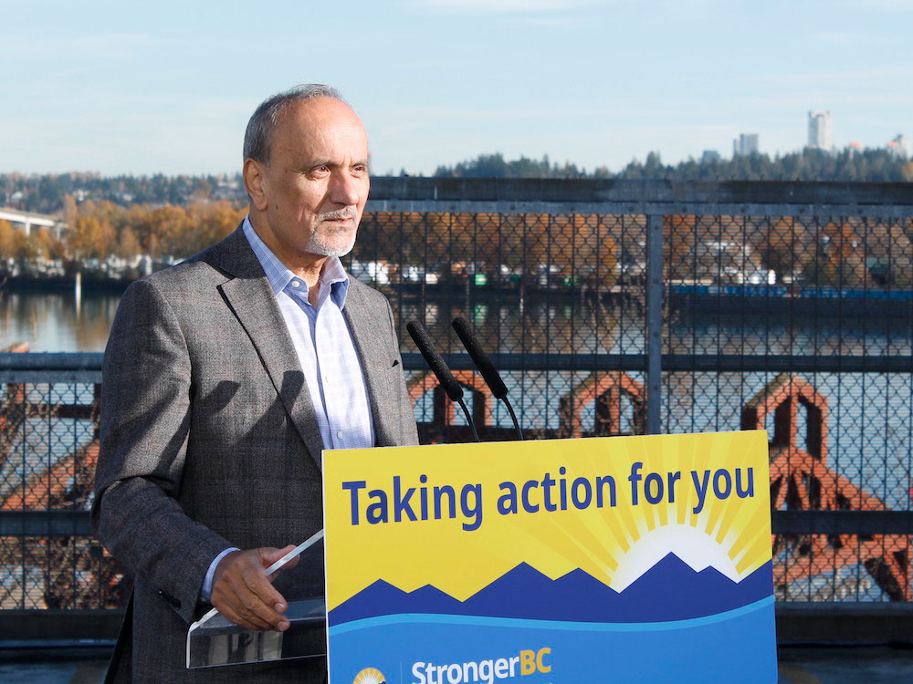 An Indo-Canadian man with grey hair and goatee stands behind a podium with a yellow sign saying 'Taking action for you.' He's wearing a grey suit and blue shirt. A river can be seen in the background.