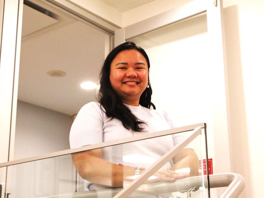 A Filipina woman with long black hair wearing a white shirt smiles at the camera on a stairway.