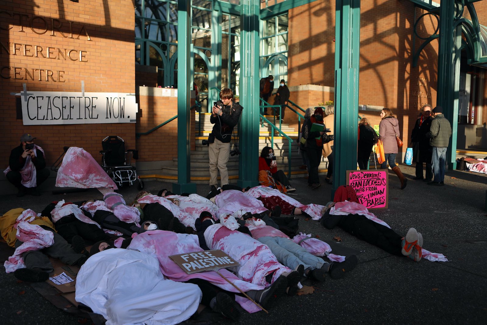 Protesters lie under sheets dyed pink and red outside the Victoria Conference Centre, with a sign saying 'Ceasefire Now' attached to a brick wall.