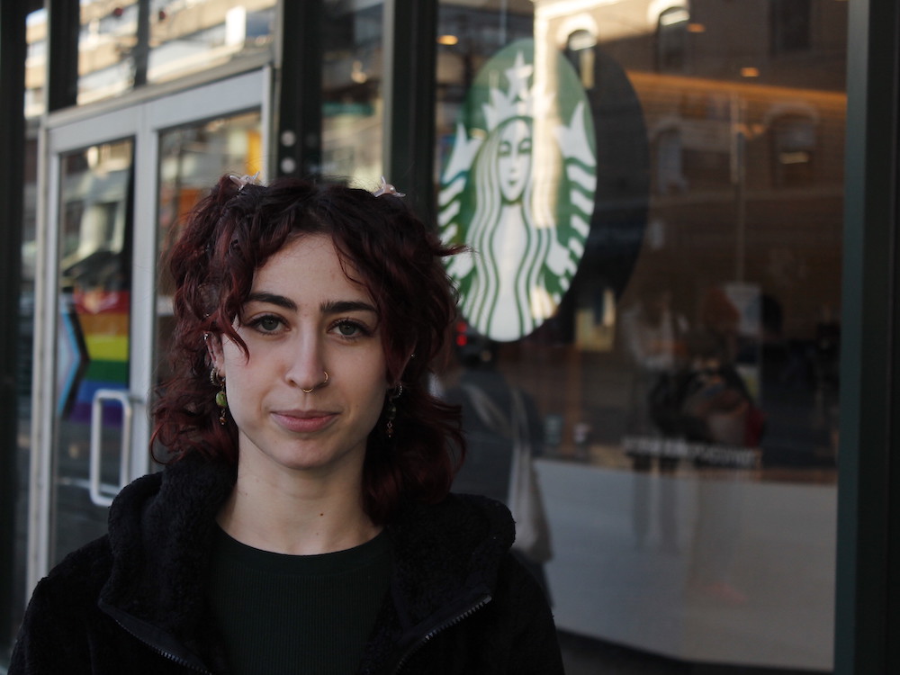 A woman with reddish hair and two nose rings, wearing a black shirt and jacket, gazes at the camera. She is standing outside a Starbucks outlet, with the logo visible beside her.