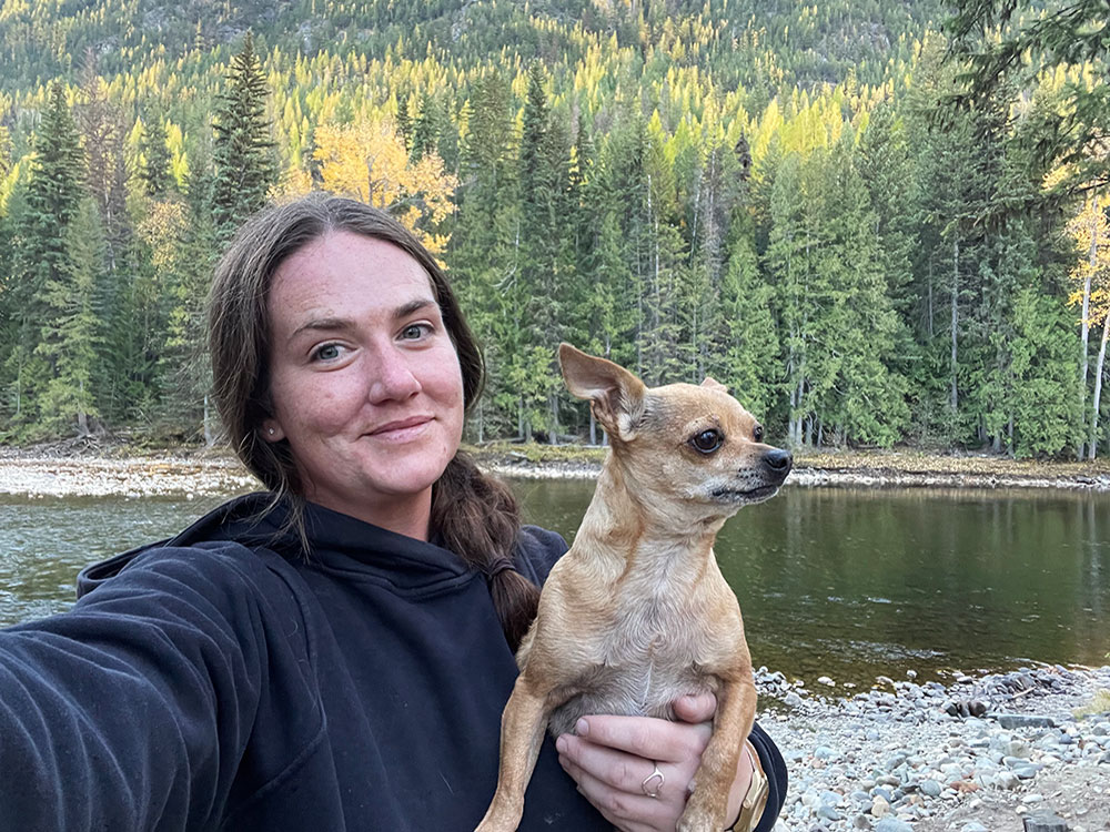 A light-skinned woman with long dark wavy hair pulled into a low ponytail poses with a small dog in front of a lake ringed with evergreens.