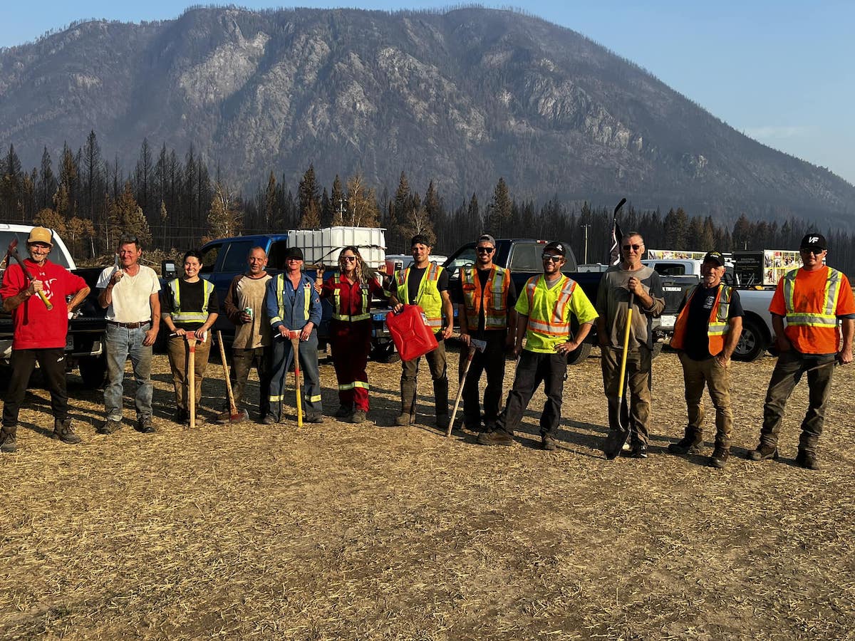 A dozen men and women, some wearing high-visibility clothing and holding tools like shovels and axes, stand in a line, smiling.