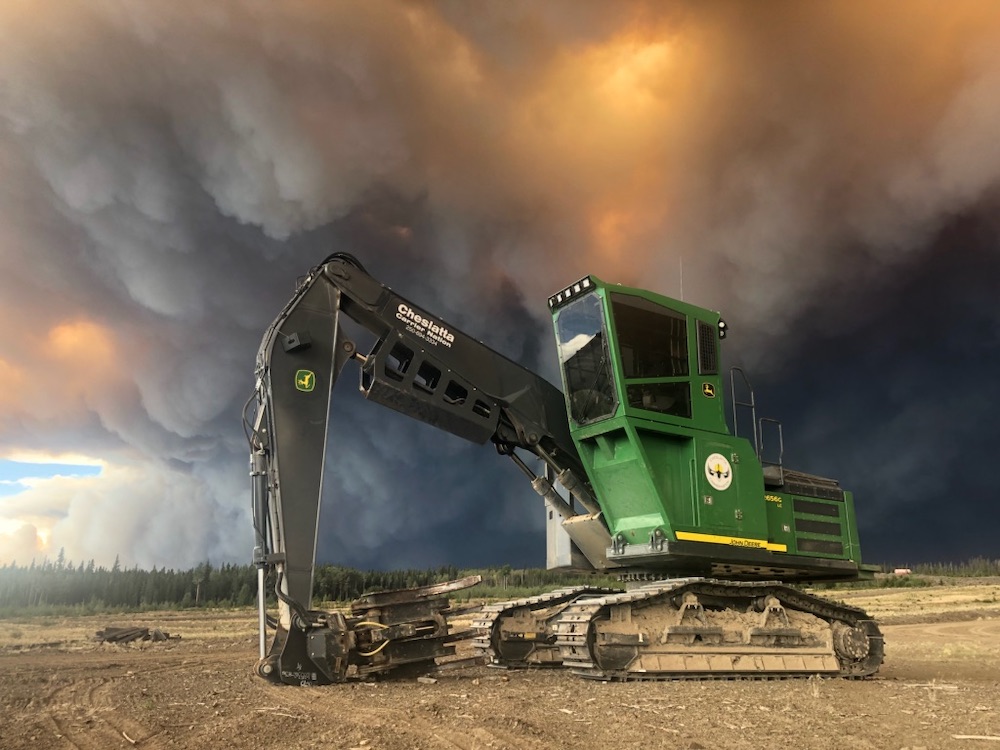 A large green vehicle on tracks sits idle in an area cleared of trees. A large, dark cloud of smoke looms behind it.