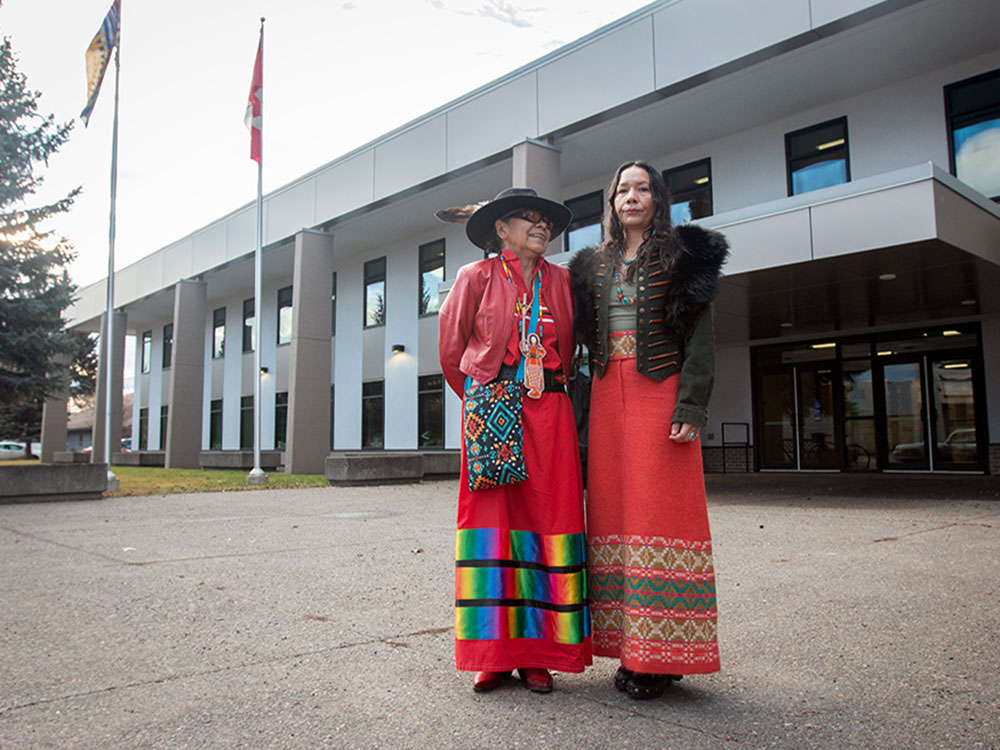 Two women, both wearing long brightly coloured skirts and one wearing a hat with an eagle feather, stand in front of a large building flying flags for Canada and British Columbia. The woman on the right looks at the camera while the woman on the left looks at her and smiles.