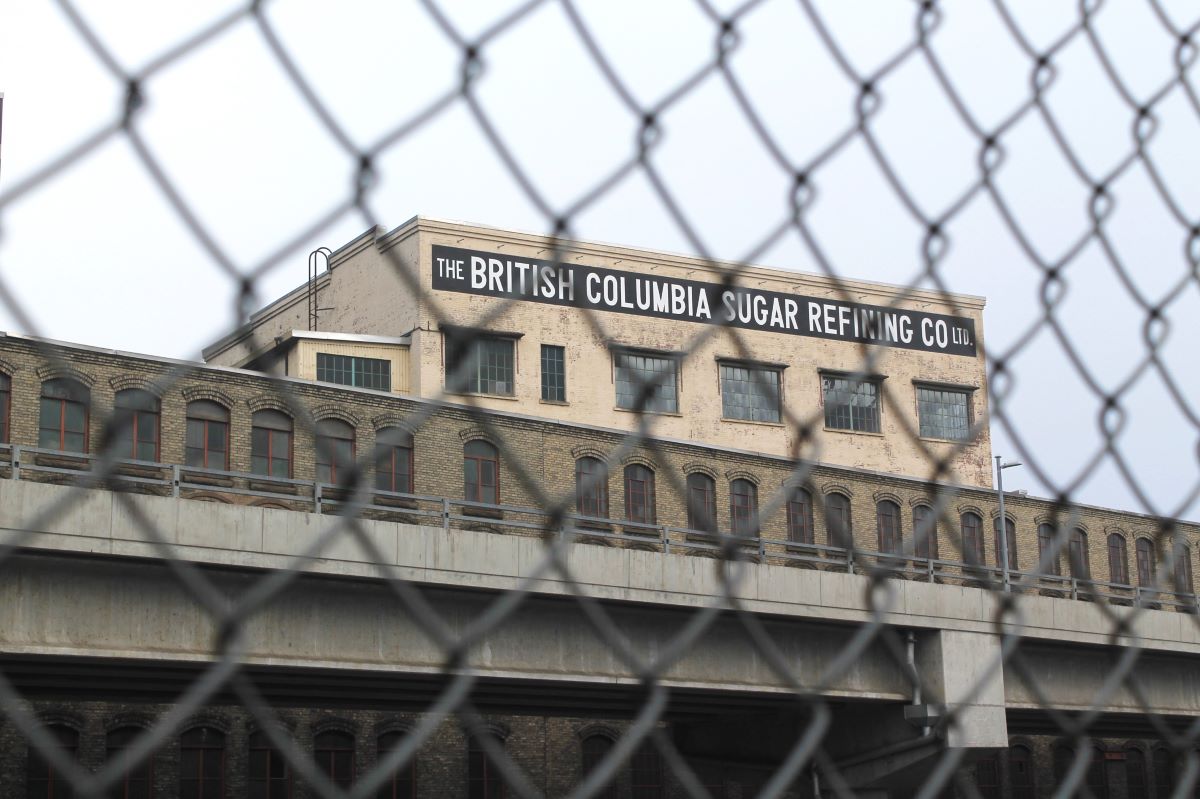 A photo taken through a chain-link fence shows a long one-storey brick industrial building. Behind it rises a taller building with a painted section on top that says 'The British Columbia Sugar Refinery Company Ltd.'
