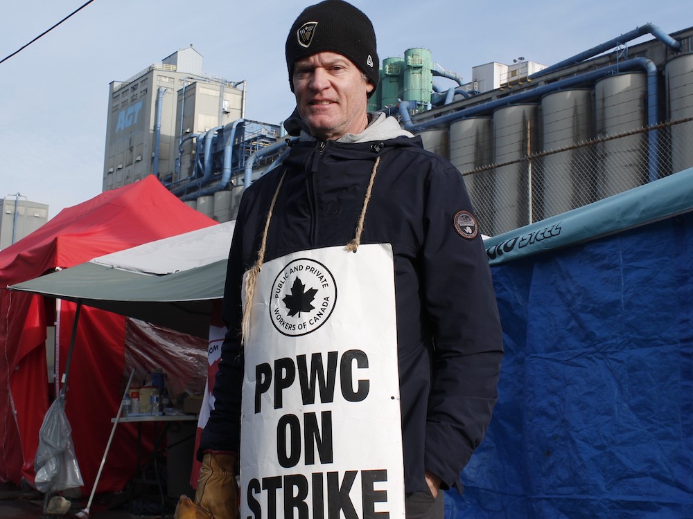 A white man wearing a black toque and black clothes looks at the camera. He has a white sign that says "PPWC ON STRIKE" and stands in front of shelters set up outside a chain-link fence.