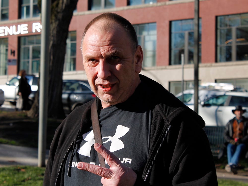 A white man with short hair and a black shirt and jacket looks at the camera. He is outside on a sunny day.