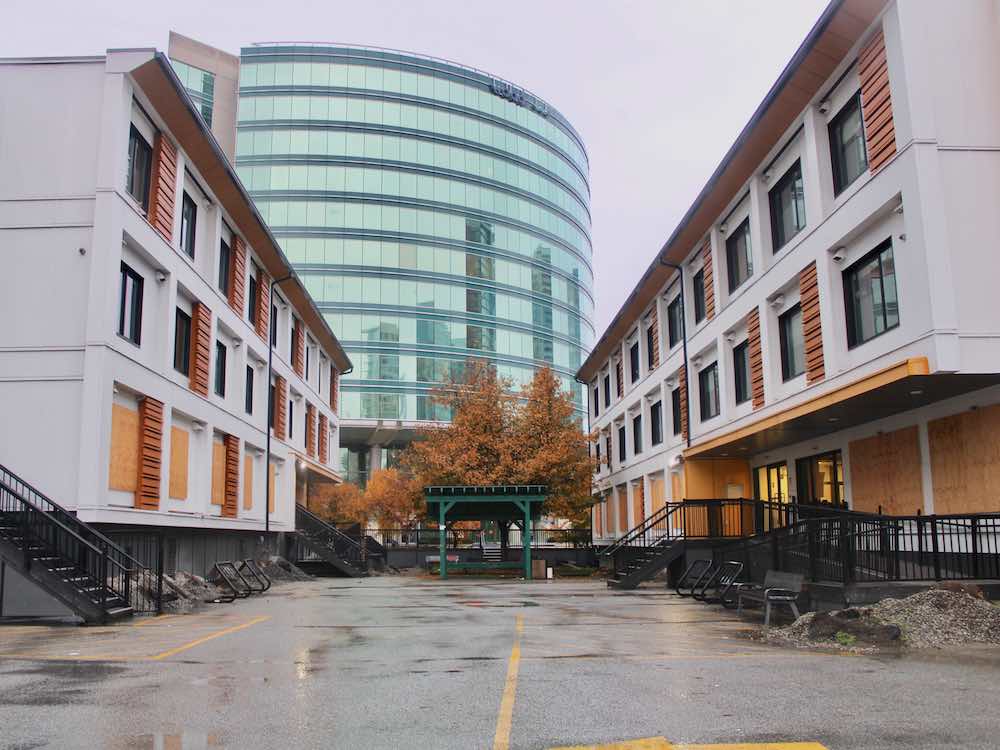 Two rows of empty three-storey modular housing face other across a rainy courtyard. They have white facades and wood accents. In the background is a curved glass building.