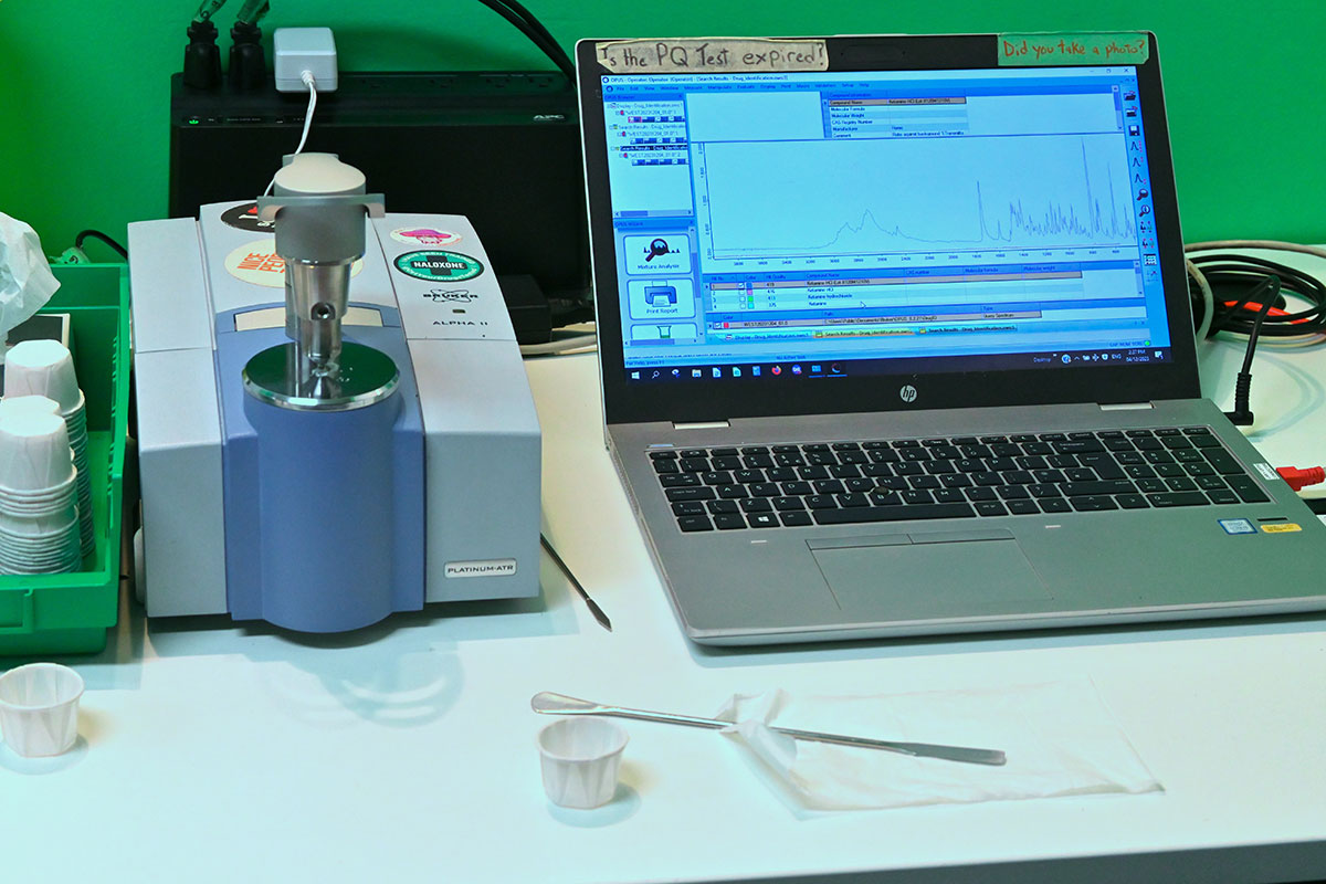 A Fourier transform infrared spectrometer on the left, and a laptop shows a drug testing result on the right.