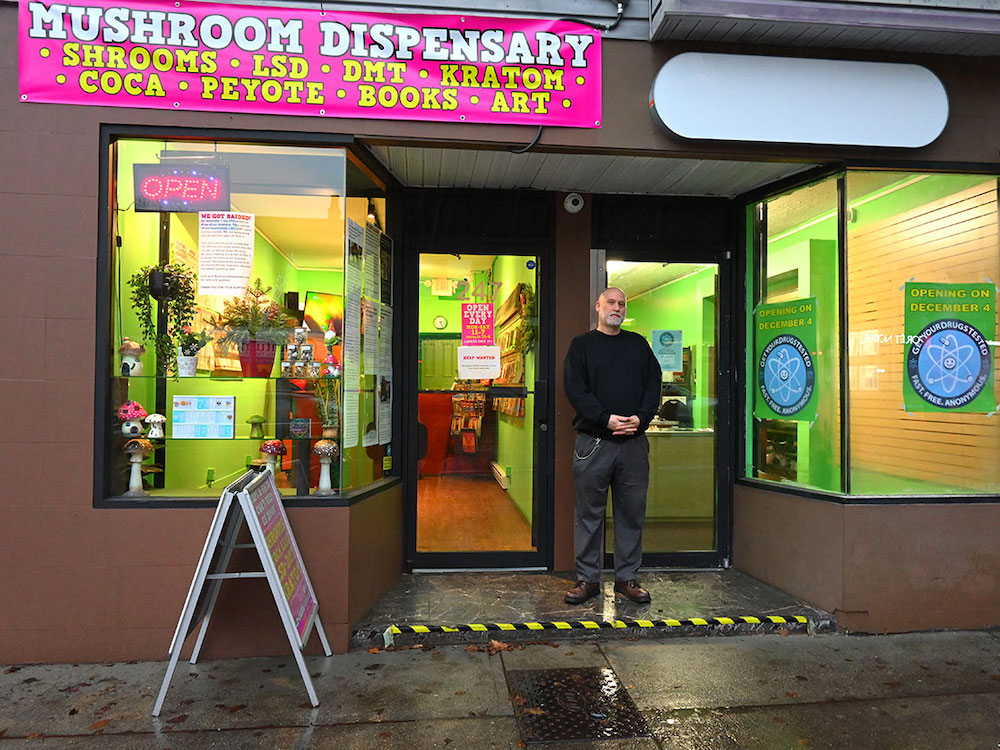 A man with light skin and a close-cropped grey beard and hair stands on the sidewalk. On one side of him is a mushroom dispensary. On the other side is the new location for Get Your Drugs Checked.