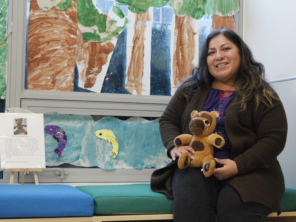 A woman with long dark hair smiles at the camera. She wears a brown sweater and purple blouse and holds a stuffie. Behind her are children's artworks.