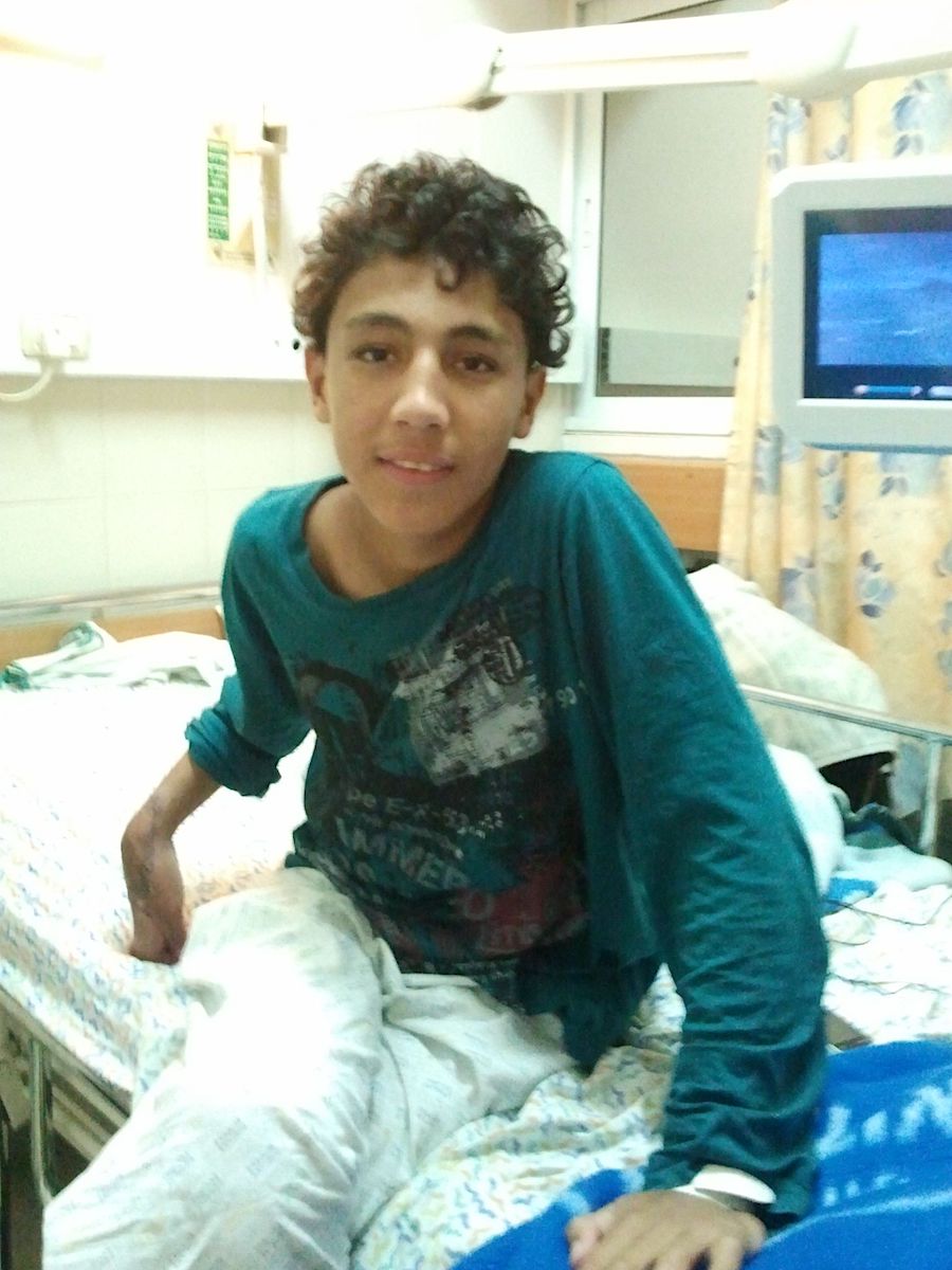 A teenage boy with dark skin tone and curly dark hair sits on a hospital bed and smiles at the camera. His right hand appears broken.