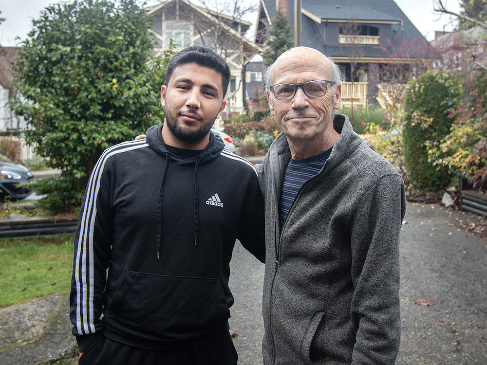 Two men stand side by side outside with shrubbery and houses behind them. The man on the left has dark skin tone and dark hair. The man on the right is older, balding and wears glasses.