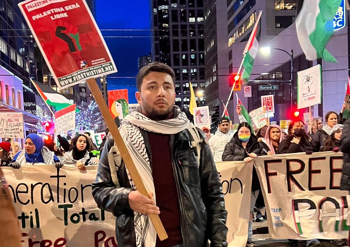 A man with dark skin tone, hair and beard and wearing a Palestinian scarf stands holding a sign in a city setting. Behind him, dozens of people stand together carrying signs, Palestinian flags and banners.