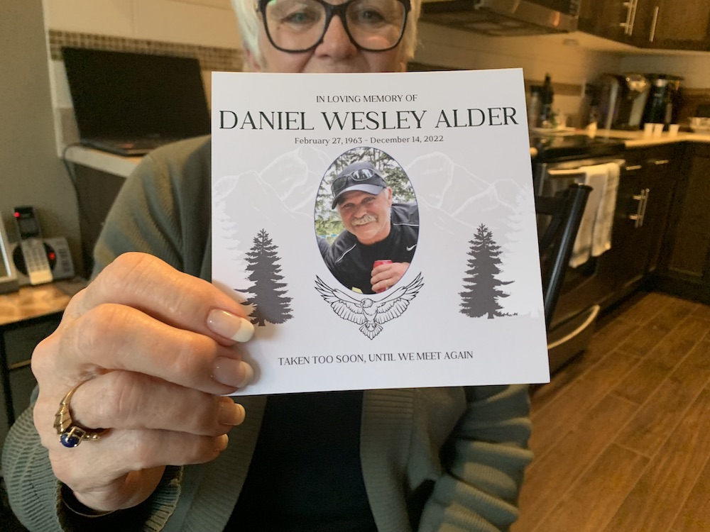 A woman with blond hair and wearing glasses, in a kitchen, holds a memorial card with a photo of a friendly-looking man wearing a ball cap.