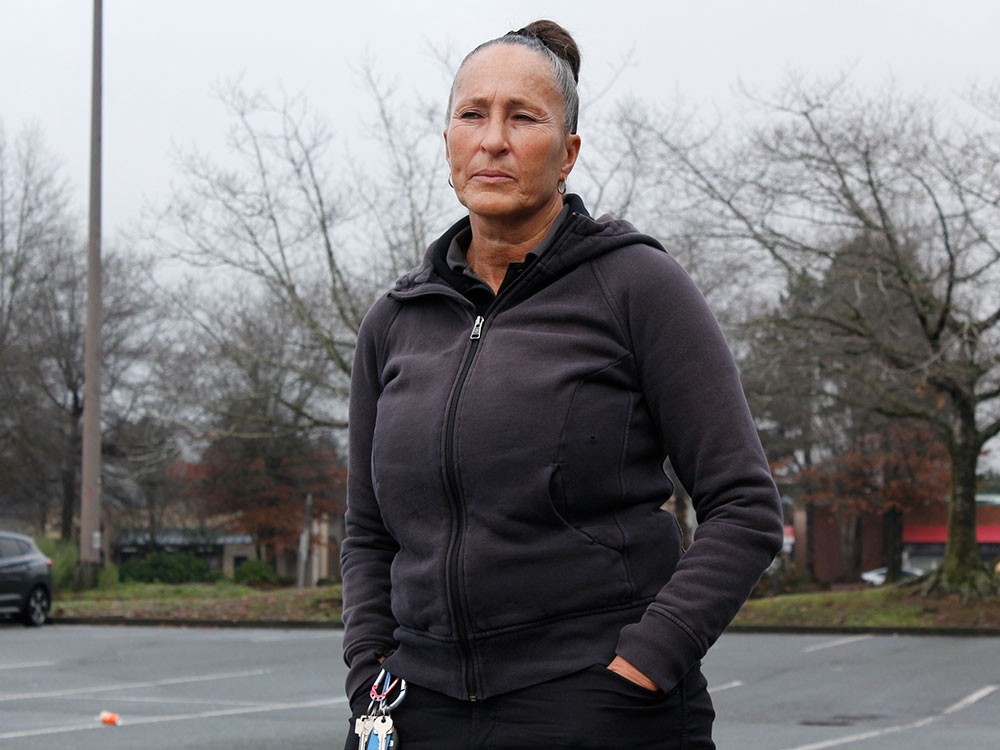 A middle-aged woman with black hair pulled back in a tight bun looks at the camera, unsmiling. She wears a black hoodie and stands in a parking lot on a grey fall day.