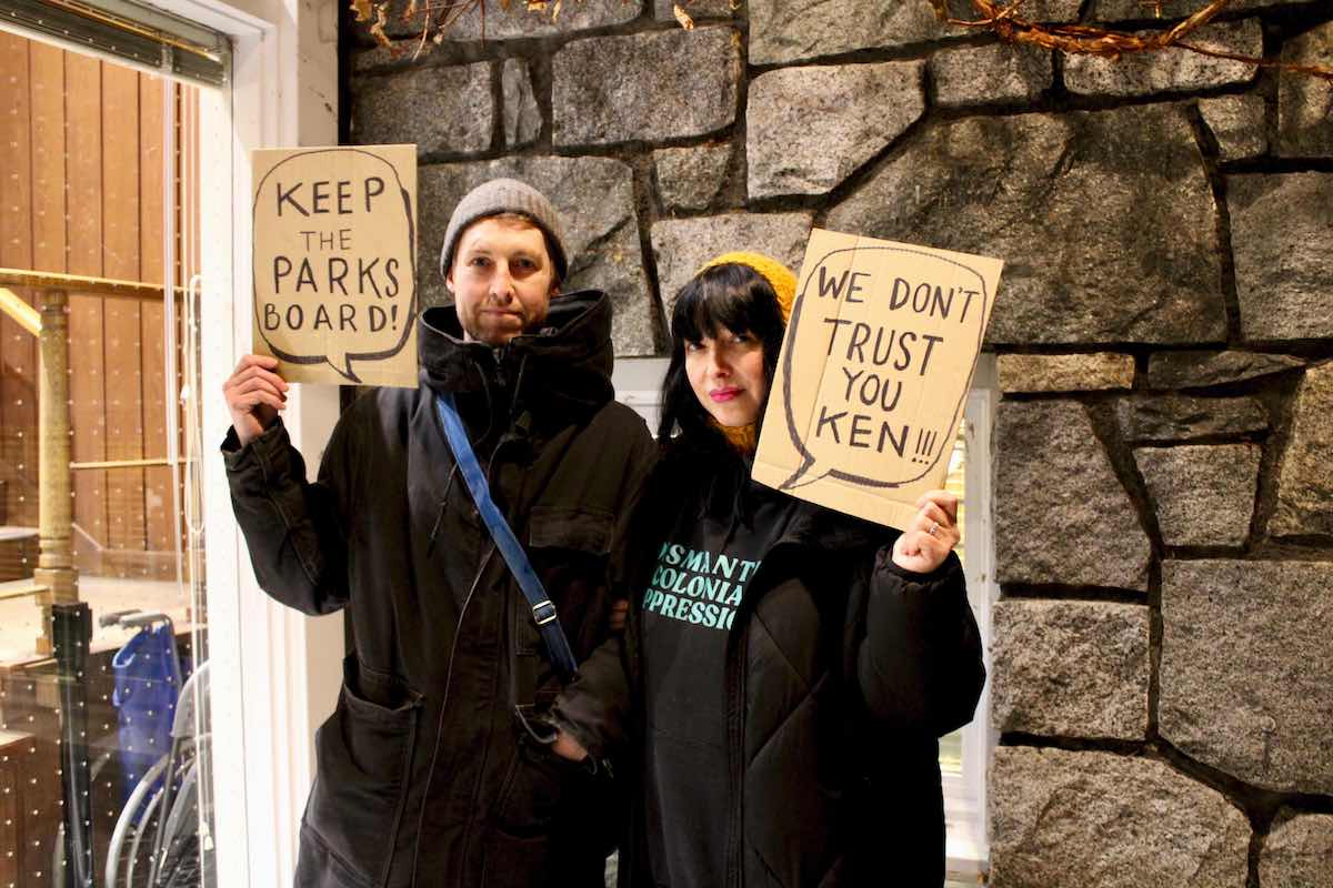 Two young people dressed in black hold cardboard signs that say 'Keep the parks board' and 'We don’t trust you Ken.'