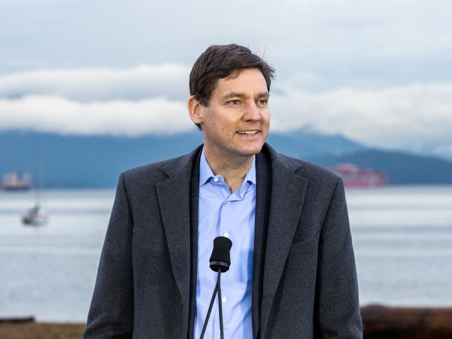 David Eby, a man with light skin tone and brown hair wearing a light blue shirt and grey jacket, stands behind a microphone, smiling and looking off to the side of the frame. Water and cloud-shrouded mountains can be seen behind him.