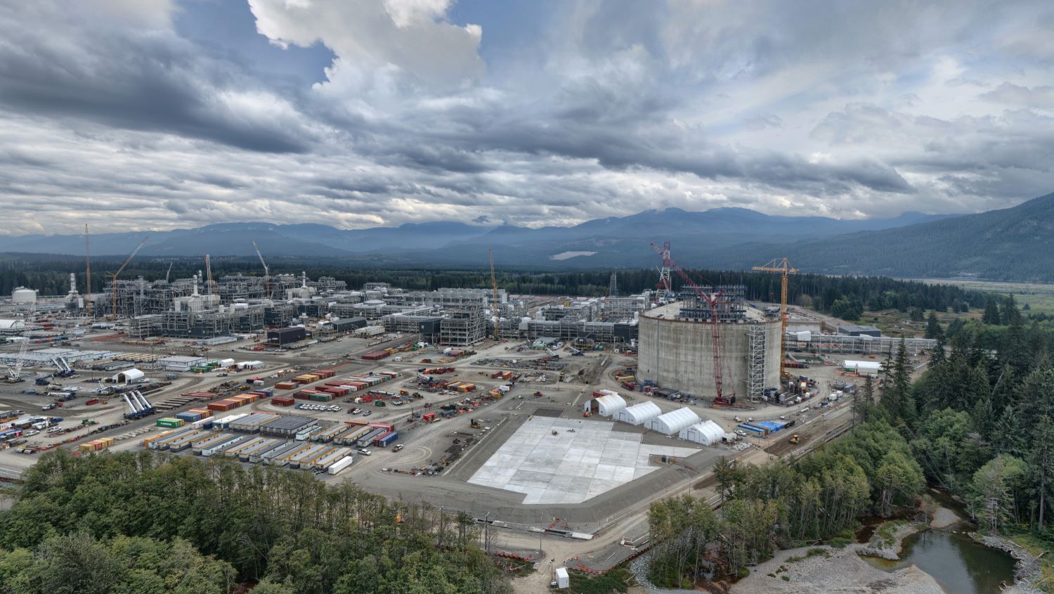 The photo shows a sprawling industrial complex under construction, with dozens of large buildings, cranes and a giant round concrete structure. It’s a cloudy day, and mountains are visible in the background.
