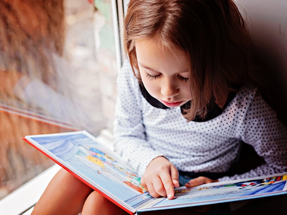 A young child with light skin and brown hair cut into a bob reads independently, her finger tracing the words in the book.
