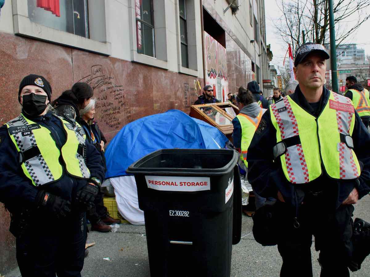 Two Vancouver police officers stand on either side of a tall black garbage can labelled 'Personal storage.' On the left, an officer with blond hair and a black toque, yellow safety vest and black medical mask looks on with their hands in their pockets. On the right, an officer with a black ball cap, yellow safety vest and black pants looks to the middle distance. Behind them is a crowd of residents and workers in neon yellow safety vests.