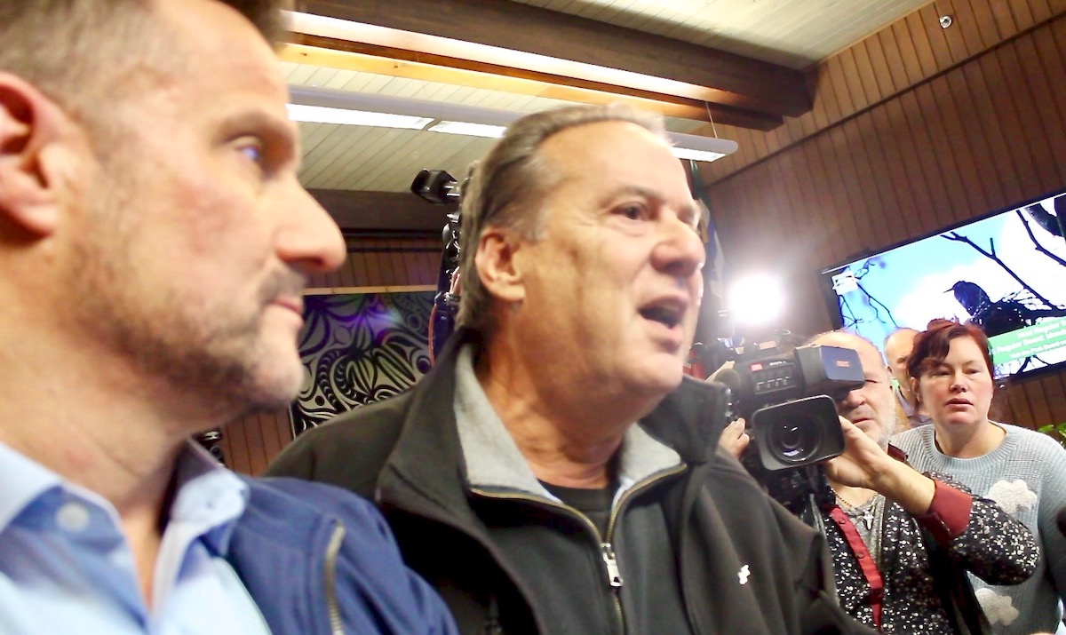 Two men crowd the centre of the frame in a heated moment. To the left, Aaron Jasper has short brown hair and stubble and is wearing a blue jacket over a light blue button-down shirt. In the middle, John Coupar is mid-speech. He has short wavy grey hair and is wearing a dark grey pullover under a black jacket. In the background is a television news camera and a crowd of people.