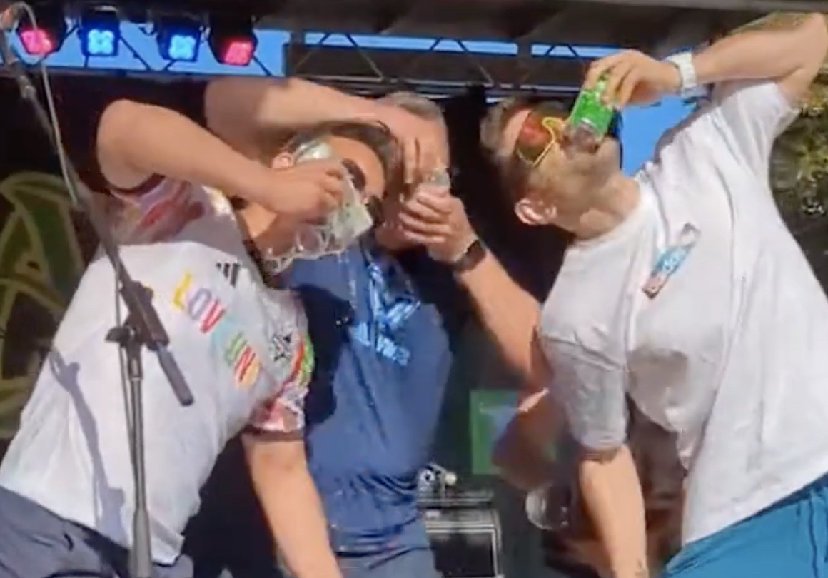 A video still depicts three men in summer clothes and oversized sunglasses holding their mouths to the open sides of cans of beer. On the left, Vancouver Mayor Ken Sim is in a white athletic shirt with rainbow adornments.