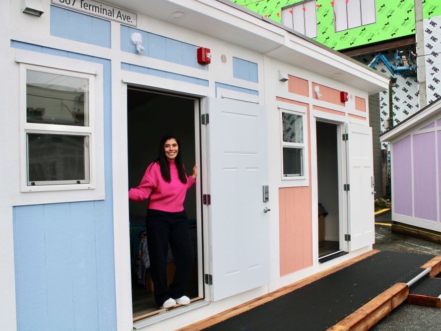 A young woman stands in the doorway of a two-unit small structure painted white, blue and rose. She is smiling and has long dark hair and wears a pink sweater, black pants and white sneakers.