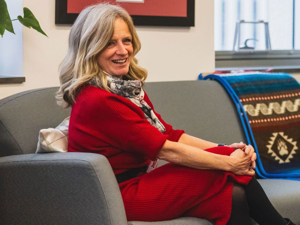 Rachel Notley, a woman with a light skin tone and medium-length blond hair, wearing a red dress and a white and black patterned scarf, sits on a grey couch at a side angle to the camera, smiling.