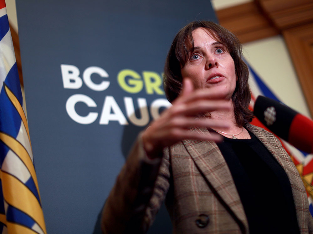 A 53-year-old white woman with shoulder-length dark brown hair gestures as she stands at a podium, wearing a black shirt and brown patterned jacket. Behind her is a B.C. flag and blue background that says 'BC Green Caucus.'