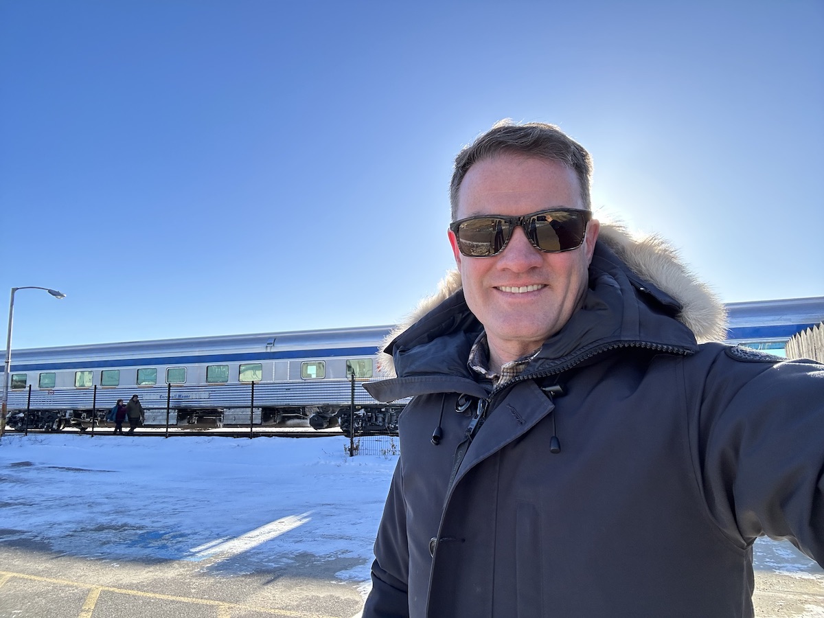 A man with light skin tone and short, dark hair and wearing a winter parka takes a selfie with blue sky and a train behind him. 