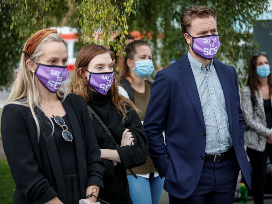 Four people stand outside wearing surgical masks that say 'Yes to SCS,' which stands for supervised consumption services.
