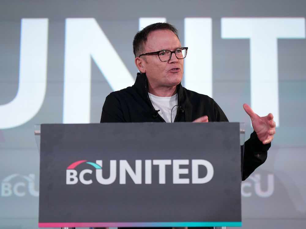 A 60-year-old white man with short dark hair and glasses stands at a podium bearing the BC United logo. He wears a white T-shirt and a black sweater with a high collar.