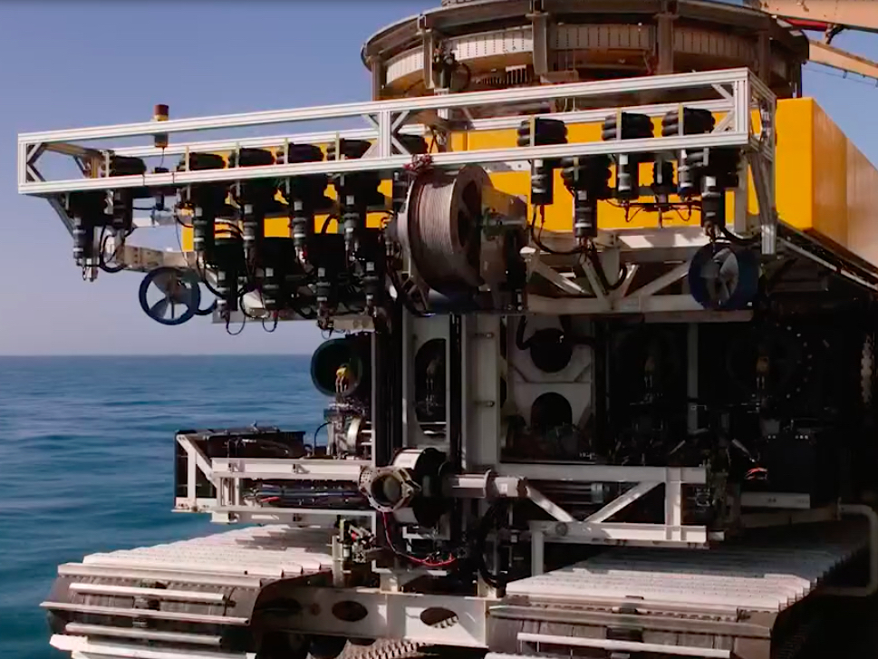 A large piece of mining equipment, equipped with silver tractor treads to travel the sea bottom, hangs over the ocean. Its superstructure is yellow and supports tubes and pumps.